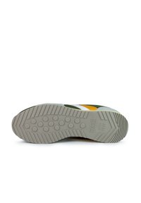 Zapatilla con suela de goma gris, parte superior en verde y amarillo, rayas blancas y logo. Superficie texturizada y suela con patrón para tracción.