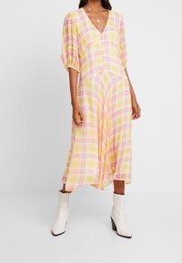 Keltainen ruudullinen midi-mekko, jossa on vaaleanpunaisia yksityiskohtia, puffhihat, V-pääntie ja napitus. Yhdistettynä valkoisiin nilkkabootseihin.