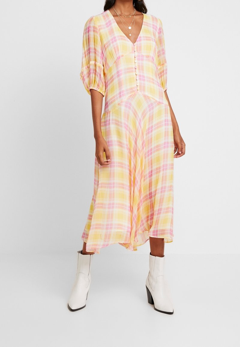 Keltainen ruudullinen midi-mekko, jossa on vaaleanpunaisia yksityiskohtia, puffhihat, V-pääntie ja napitus. Yhdistettynä valkoisiin nilkkabootseihin.