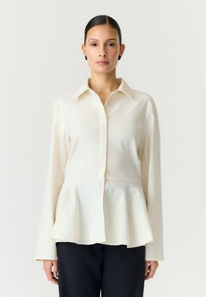 Femme aux cheveux foncés avec raie au milieu, portant des boucles d'oreilles noires, une chemise blanche à manches longues avec un ourlet péplum et un pantalon foncé.