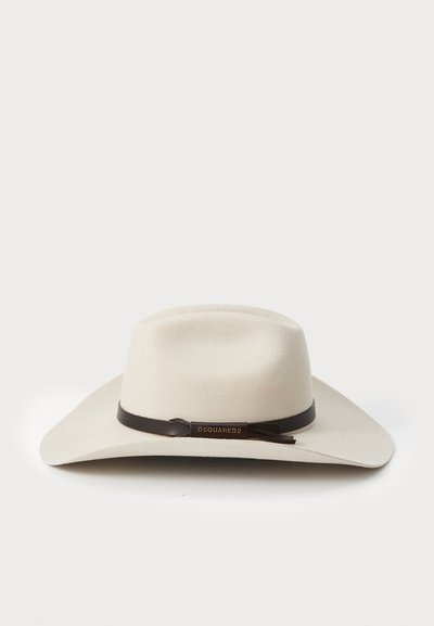 Chapéu de aba larga em feltro creme com uma faixa de couro castanho escuro e um detalhe em logotipo dourado. Apresenta uma forma clássica e estruturada.