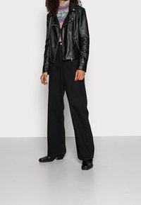 Schwarze Leder-Bikerjacke mit silbernen Reißverschlüssen, kombiniert mit einem mehrfarbigen, gemusterten Oberteil und weiten schwarzen Hosen. Schwarze Stiefeletten mit Absatz.