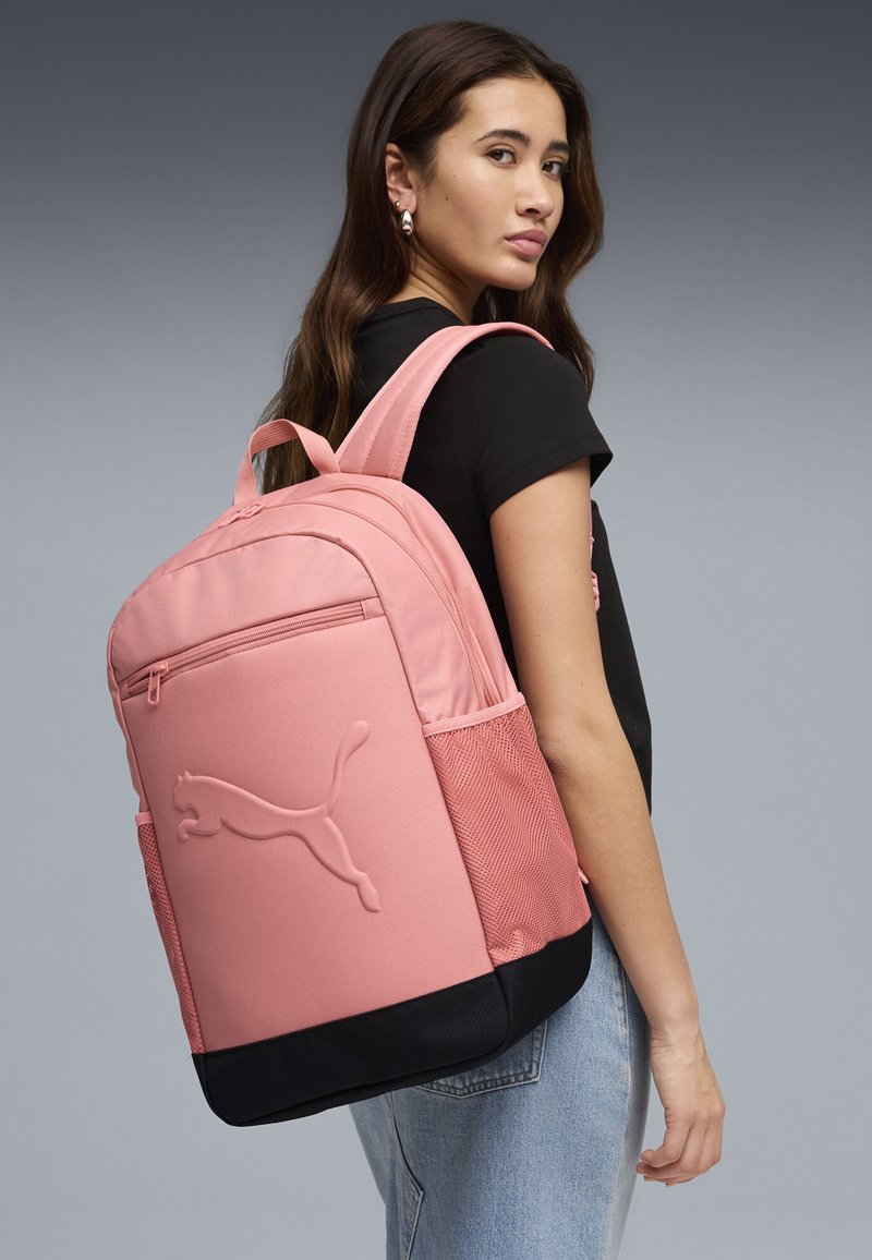 Jeune femme portant un sac à dos Puma rose avec une poche zippée à l'avant et une base noire, vêtue d'une chemise noire et d'un jean bleu, vue de côté.