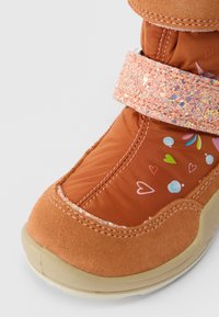 Gyerek narancssárga téli csizma csillogó rózsaszín pánttal, szív- és buborékmintákkal, bézs talppal fehér háttér előtt.