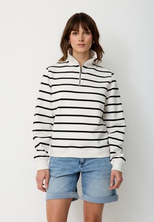 Frau trägt einen weißen Langarm-Pullover mit schwarzen horizontalen Streifen und einem Frontreißverschluss, kombiniert mit hellblauen, hochgekrempelten Jeansshorts.