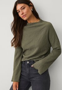 Olivgrünes, strukturiertes Langarmoberteil mit hohem Kragen und ausladenden Ärmeln, kombiniert mit dunkelblauen, hoch taillierten Jeans.