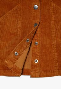 Close-up van een roestkleurige corduroy jas met zilveren drukknoopjes, waarvan sommige zijn losgemaakt om de bijpassende stofvoering eronder te onthullen.