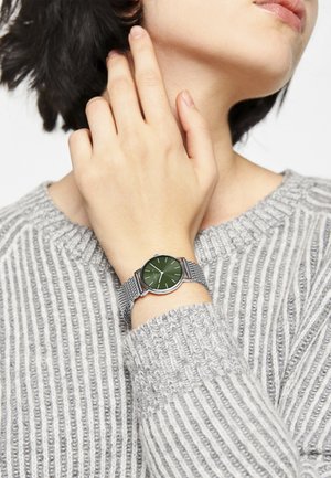 Green watch with a round face and silver mesh band, worn on a wrist. The wearer has a gray, ribbed sweater. Simple, minimalistic design.