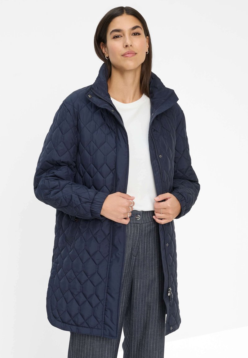 Marineblå quilted lang frakke med høj krave, trykknapper og sidelommer. Glat tekstur med et diamantmønster. Båret over en hvid top.