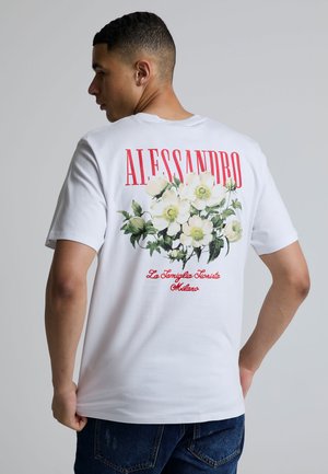 Man draagt een wit T-shirt met rode tekst "ALESSANDRO", een afbeelding van witte bloemen en rode cursieve tekst "La Famiglia Fiorista Milano" op de achterkant.
