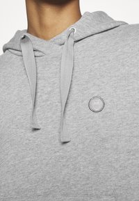 Haut à capuche gris en tissu doux avec une poche kangourou, un cordon de serrage à la capuche et un logo circulaire de hibou brodé sur la poitrine.
