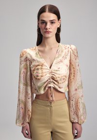 Langärmlige, cropped Bluse in hellbeige mit Blumen- und Blattmustern, mit gerafftem Vorderteil und Bändern. Weiche, fließende Stoffstruktur.