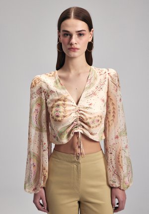 Blusa a maniche lunghe e corta in beige chiaro con motivi floreali e foglie, caratterizzata da un ventre arricciato e lacci. Tessuto morbido e fluido.