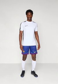 Nike spordirõivaste komplekt, mis sisaldab valget lühikeste varrukatega särki mustade detailidega ja siniseid lühikesi pükse logoga. Komplekteeritud mustade kingadega.