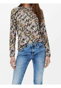 Blusa floreale con maniche lunghe, scollatura arricciata e motivi colorati nei toni di viola, rosa e giallo. Abbinata a jeans azzurri chiari.