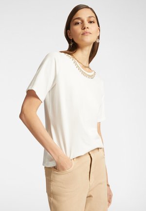 T-shirt bianco a maniche corte con scollatura decorativa e accenti dorati, abbinato a pantaloni beige chiaro, che evidenziano una vestibilità comoda e uno stile casual.