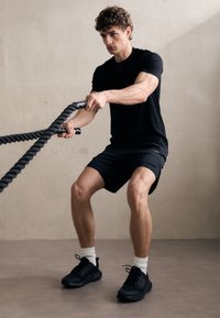 Camisa deportiva negra, pantalones cortos negros y zapatillas negras. Persona utilizando cuerdas de batalla gruesas, involucrada en una rutina de ejercicio, con un fondo neutro.