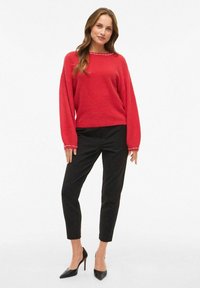 Maglione rosso con maniche lunghe e scollo rotondo, con dettagli decorativi. Abbinato a pantaloni aderenti neri e tacchi a punta neri.