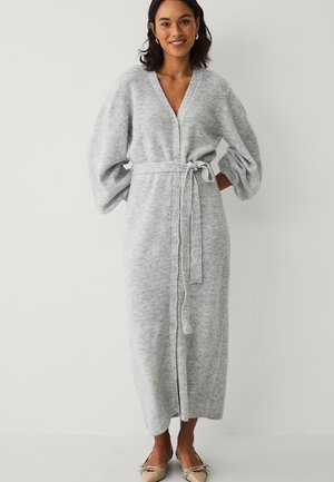 Vrouw draagt een lange, lichtgrijze gebreide jurk met knopen, pofmouwen en een ceintuur in de taille, gecombineerd met beige spitse ballerina's.