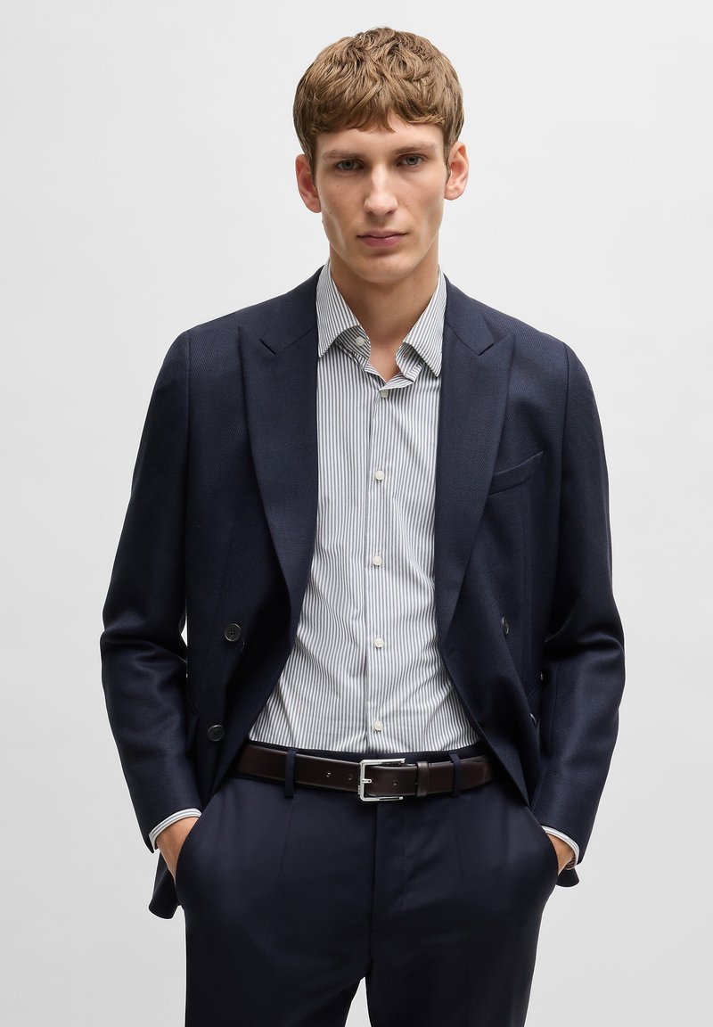 Blazer bleu marine à double boutonnage, chemise à rayures ajustée, ceinture sombre, pantalon sur mesure, tissu lisse, design structuré et boutons proéminents.