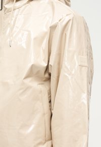 Μπεζ γυαλιστερό αδιάβροχο με κουκούλα, πλαϊνή τσέπη και μικρό ορθογώνιο λογότυπο στο πάνω μέρος του μπράτσου.