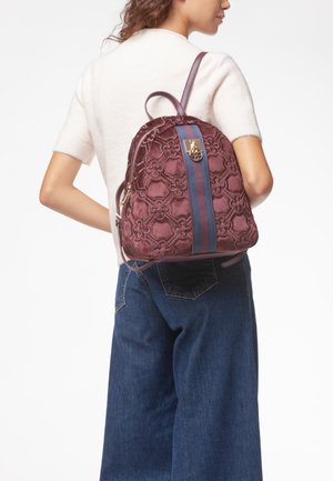 Donna che indossa una maglietta bianca a maniche corte e jeans blu porta uno zaino bordeaux strutturato con strisce blu e rosse sulla schiena.
