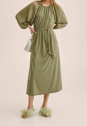 Midi-jurk in olijfgroen met geplooide ballonmouwen, een getailleerde taille en bijpassende groene platte sandalen met een zachte uitstraling. Geweven witte handtas als accessoire.