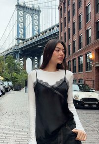 Donna con lunghi capelli scuri che indossa un top di pizzo nero sopra una maglietta bianca a maniche lunghe, in piedi su una strada acciottolata vicino a un ponte e a edifici in mattoni.