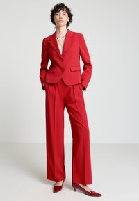 Roter, maßgeschneiderter Anzug mit einem taillierten Blazer und weiten Hosen. Hergestellt aus glattem Stoff; verfügt über zwei Knöpfe und Vordertaschen. Glänzende rote Absatzschuhe.