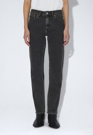 Persoon draagt zwarte high waist denim jeans met rechte pijpen, gecombineerd met zwarte enkellaarzen en een wit ingestopt overhemd.
