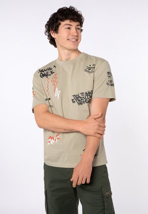 Junger Mann lächelnd in beigefarbenem Grafik-T-Shirt mit schwarzem und orangefarbenem Text und dunkelgrüner Cargohose, stehend vor schlichtem Hintergrund.