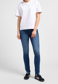 Vit bomull T-shirt med korta ärmar och en bröstficka, tillsammans med blå denimjeans och svarta sneakers med vita detaljer.