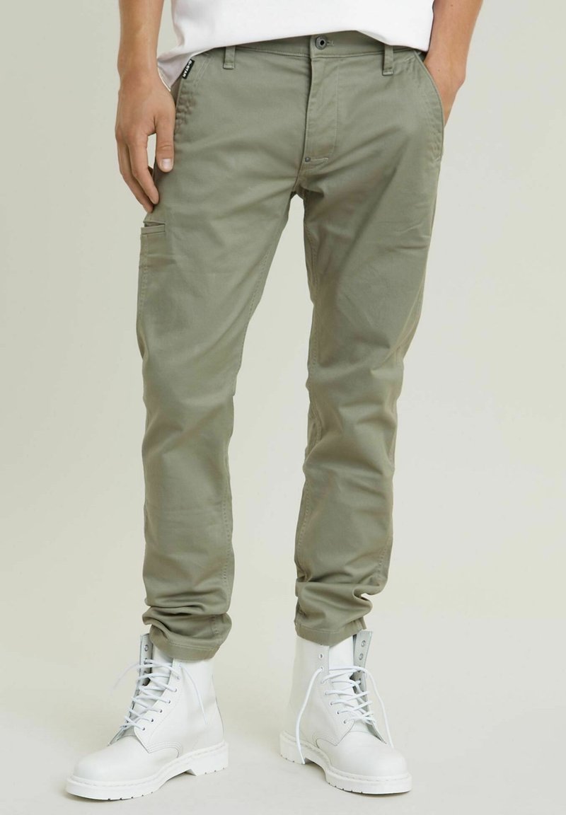 Partie inférieure du corps d'une personne portant un pantalon cargo vert olive et des bottines blanches à lacets, debout les mains dans les poches.