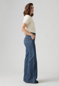 Wide-leg blue denim jeans with a high waist, featuring a five-pocket design and contrast stitching. Worn with a cream short-sleeve top.