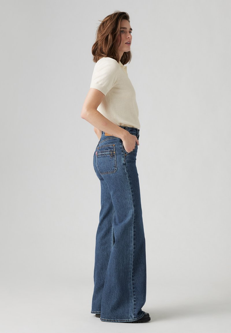 Jeans en denim bleu à jambes larges avec une taille haute, dotés d'un design à cinq poches et de coutures contrastées. Portés avec un haut crème à manches courtes.