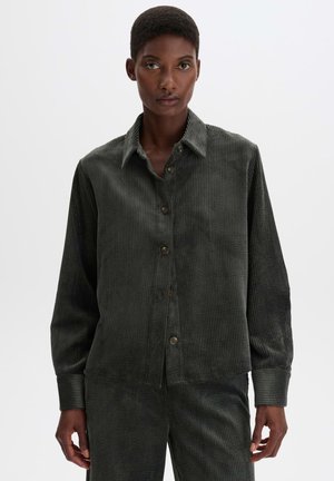 Donna che indossa una camicia in velluto a coste verde scuro abbottonata e pantaloni coordinati, in piedi davanti a uno sfondo chiaro e uniforme.