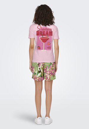 Persoon met een roze "Strawberry Dream" T-shirt met een afbeelding van een aardbeiendrank, groene en paarse bloemige korte broek en witte sneakers, van achteren gezien.