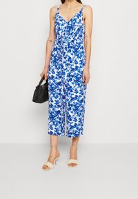 Donna che indossa una jumpsuit senza maniche blu e bianca con motivi floreali, sandali con tacco intrecciati beige, tiene in mano una piccola borsa nera.