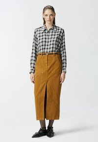 Chemise à carreaux noir et blanc avec boutons, associée à une jupe marron jusqu'aux genoux avec une fente à l'avant. Des chaussures noires à lanières complètent la tenue.