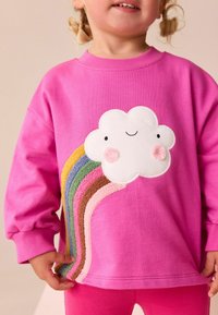 Fuchsia Sweatshirt mit einem lächelnden Wolken- und strukturierten Regenbogen-Design in verschiedenen Farben. Lange Ärmel und gerippte Bündchen. Weicher Stoff.
