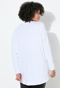 Vrouw met krullend haar, gekleed in een losse witte lange mouwen shirt, die gedeeltelijk wegdraait en over haar rechterschouder kijkt.