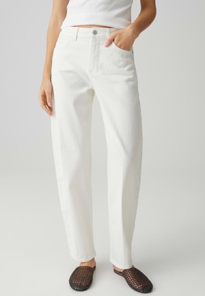 Donna che indossa jeans bianchi a gamba dritta, maglia bianca infilata e scarpe nere intrecciate senza lacci, in piedi con una mano in tasca.