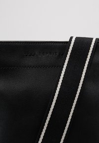 Czarna skórzana torba z fakturowanym wykończeniem i paskiem z płótna w szare paski, z wytłoczonym nazwiskiem marki na powierzchni.