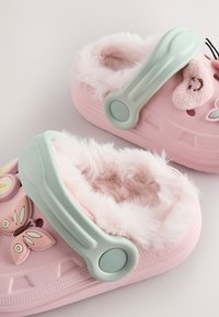 Rosa træsko med en blød, fluffy indvendig foring, grønne stropper og dekorative sommerfugle- og smilende ansigtsdetaljer på siderne.