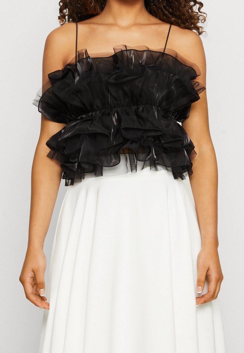 Vrouw in een zwarte, geplooide transparante crop top met dunne bandjes en een hooggesneden witte rok, met de armen ontspannen naast haar lichaam.