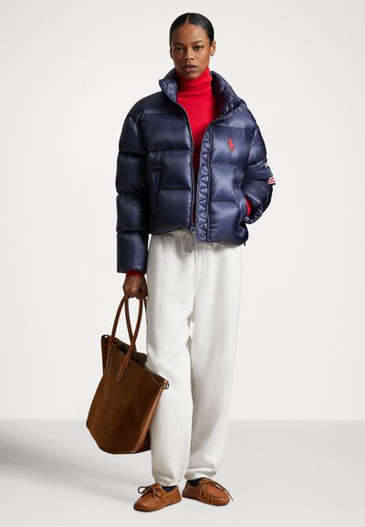 Navy puffer jacket with a high collar and red logo, paired with a red turtleneck, white sweatpants, and brown suede tote bag and shoes.