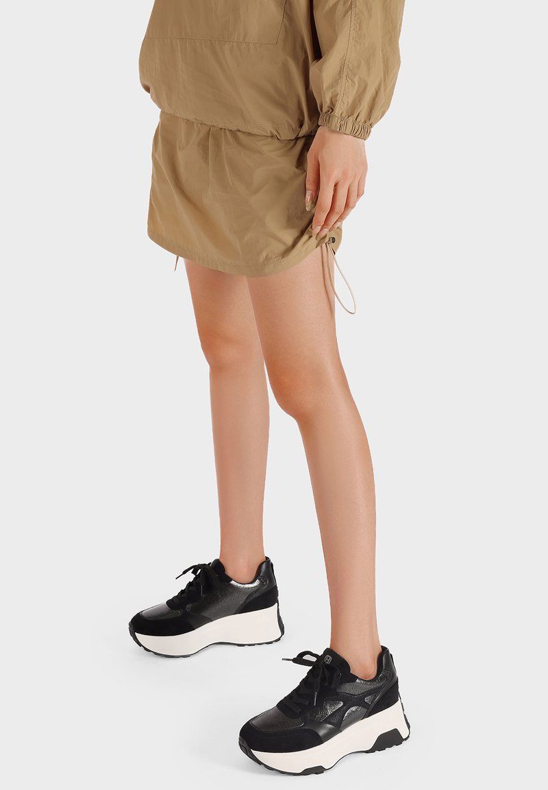 Sneakers platform bianco e nero con finitura lucida e tomaia testurizzata, abbinati a un vestito khaki con coulisse.