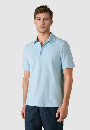 Young man wearing a light blue short-sleeve polo shirt and dark navy pants, standing against a plain light gray background.