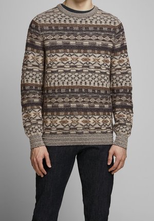 Homme portant un pull en tricot à motifs marron, beige et noir avec col rond et un jean foncé, debout devant un fond gris uni.