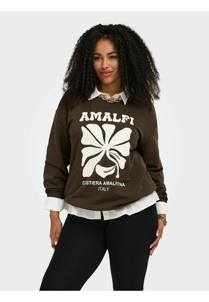 Frau mit lockigem Haar, die einen braunen Pullover mit der Aufschrift „Amalfi Costiera Amalfitana Italy“ über einem weißen Hemd und schwarzen Hosen trägt.